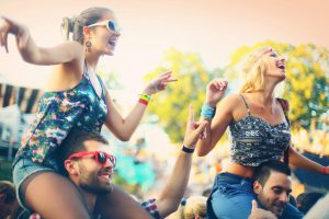 Group of young adults at a concert.Two men carrying their girls on shouldersGirls are ecstatic and having big fun and guys not so much.It's summer sunset.Large crowd in background,blurry.